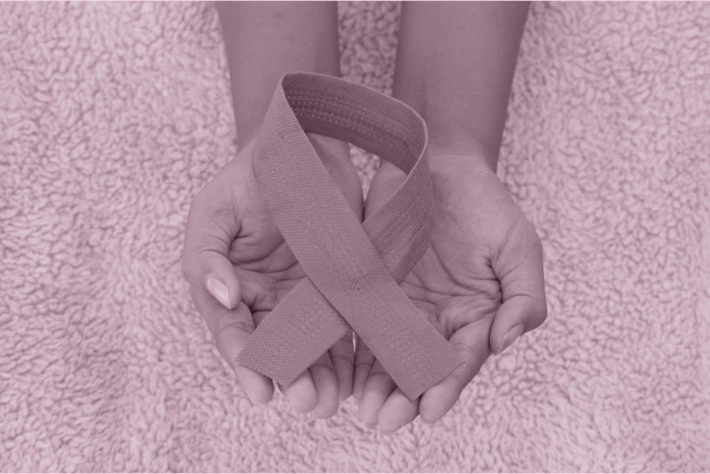 breast cancer awareness ribbon held by two cupped hands on a fleece blanket. The image is all a soft pink.